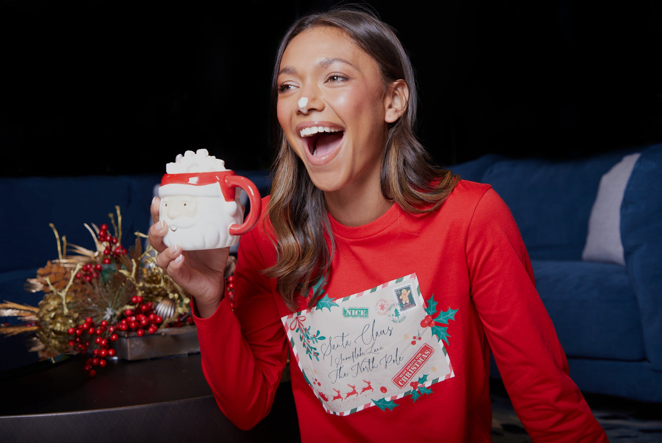 Woman in a red Christmas-themed shirt holding a white mug with a red handle, laughing.